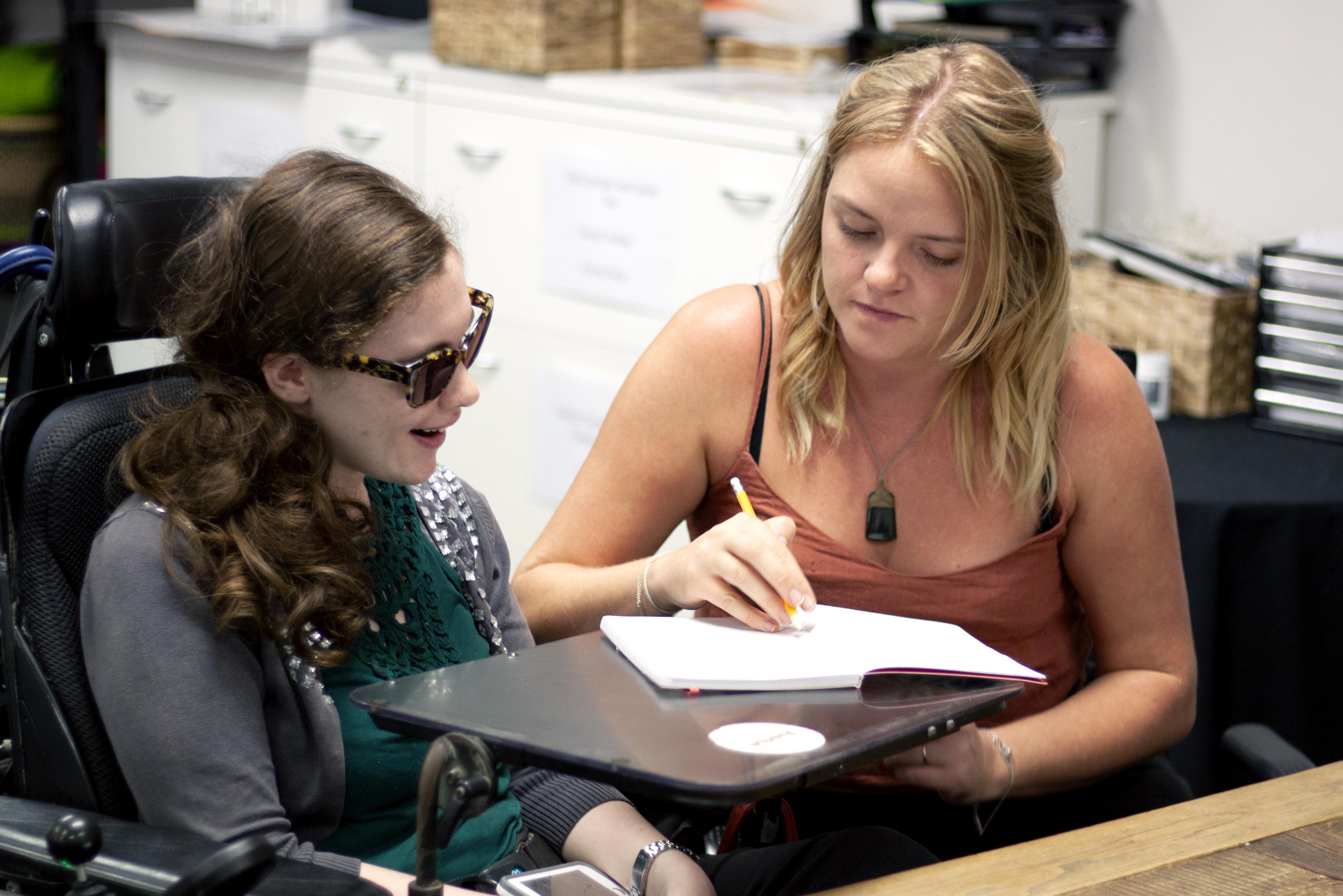 Avenue participant in wheelchair working at computer with Avenue support worker next to her. Both smiling and looking at a notebook.