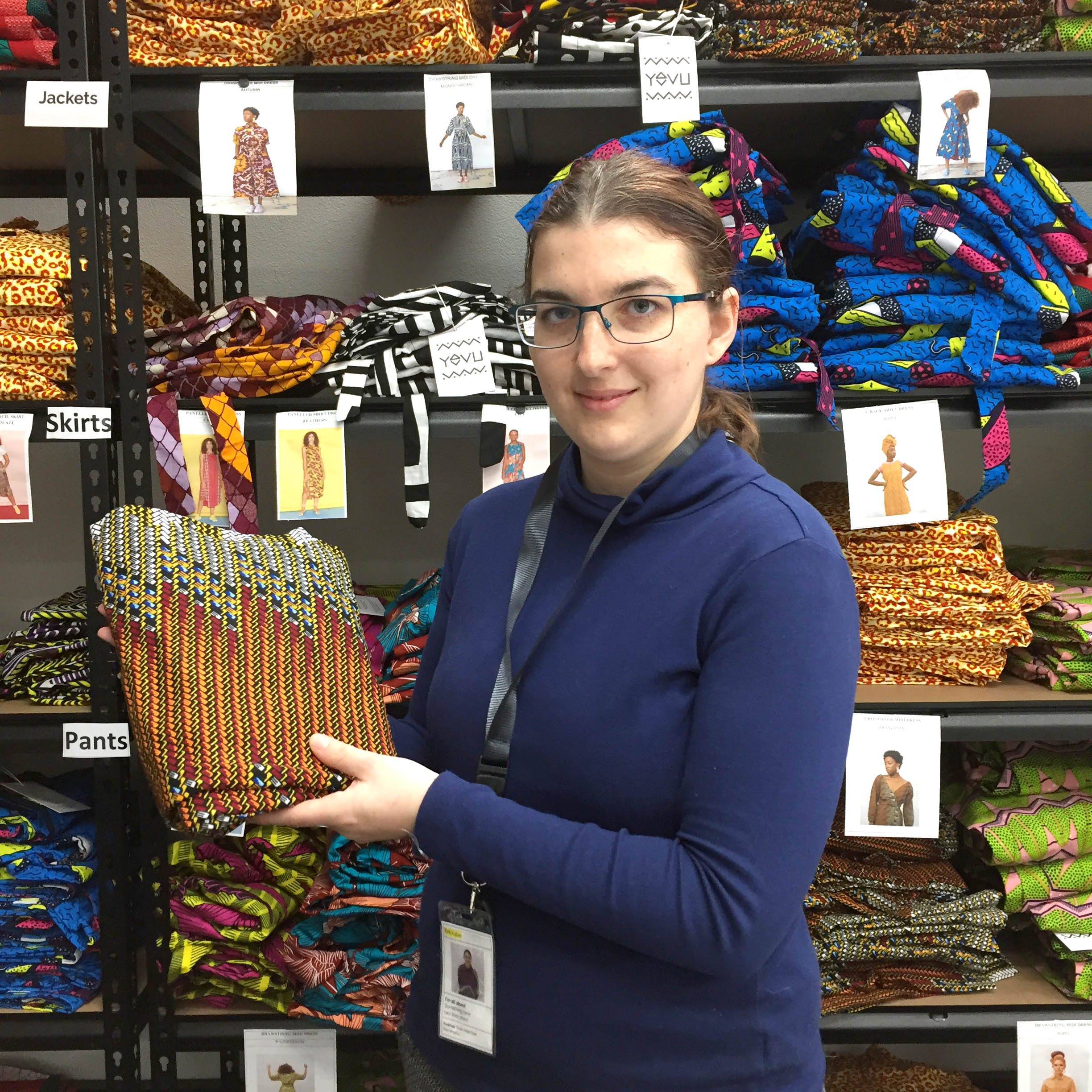 katie is wearing glasses and a navy long sleeve top. She is in the YEVU order fulfilment room in Avenue Parramatta and is holding some YEVU clothing.