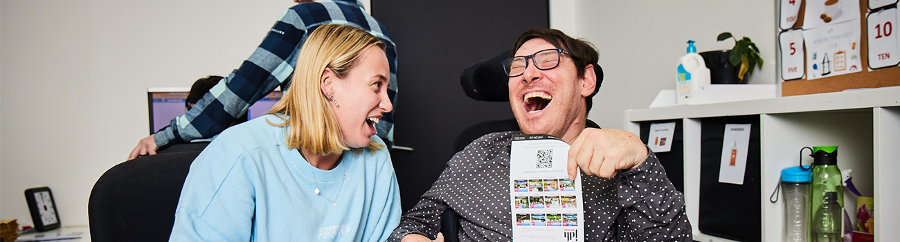 Avenue Frenchs Forest participant in the Flyers team is laughing next to his support worker, whilst holding some flyers