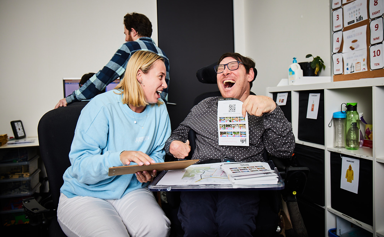 Avenue Frenchs Forest participant in the Flyers team is laughing next to his support worker, whilst holding some flyers