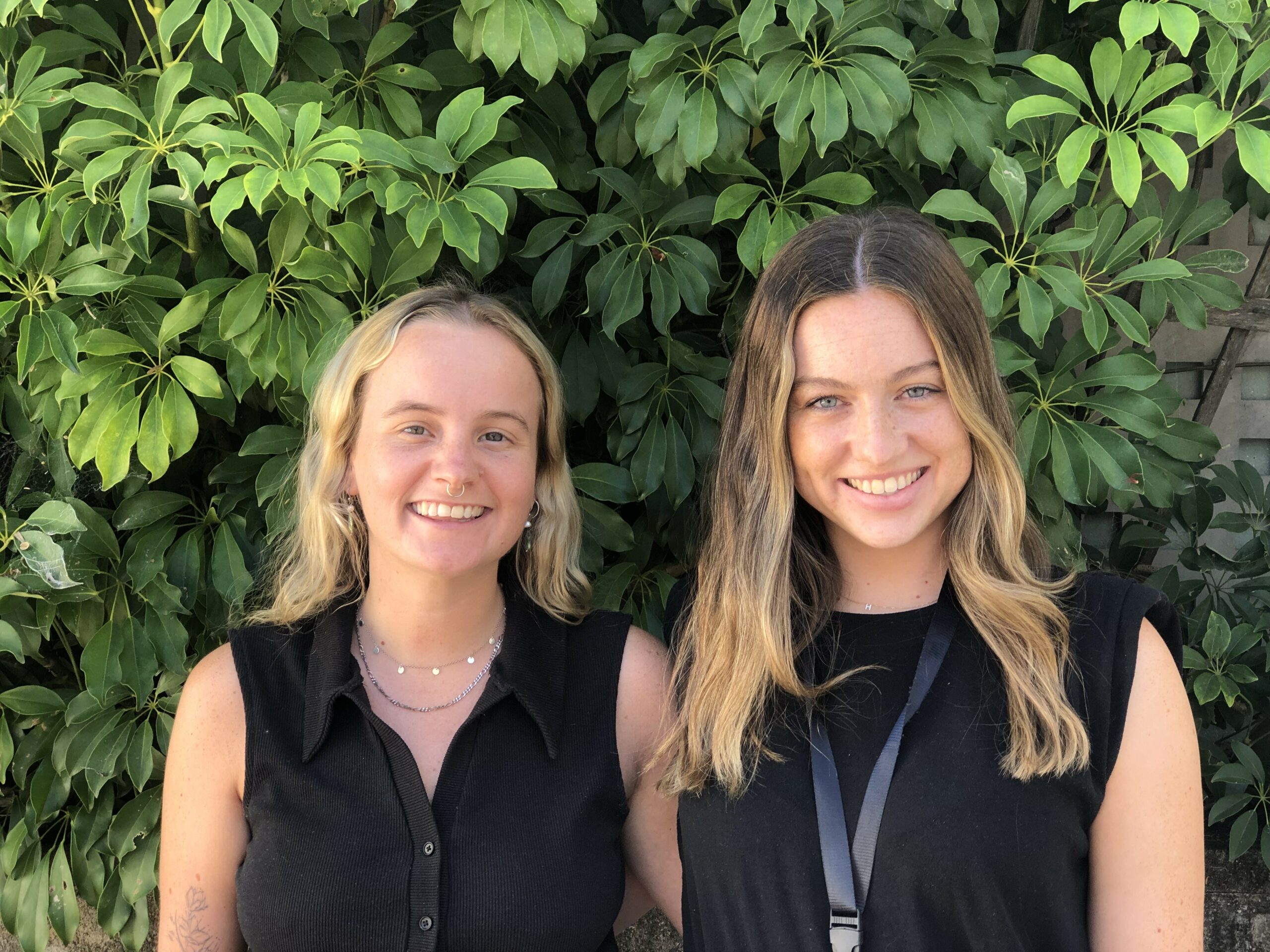 Alex and Harriet are standing in front of some trees, smiling at the camera. Alex (left) has long blonde hair and is wearing a black top. Harriet has a long ballyage and is also wearing a black top.