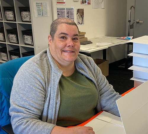 Julie is sitting at a desk wearing a green top and grey cardigan. She is folding boxes in the order fulfilment room in Avenue Newcastle