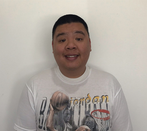 Sing is standing against a white wall, wearing a white t-shirt, smiling at the camera