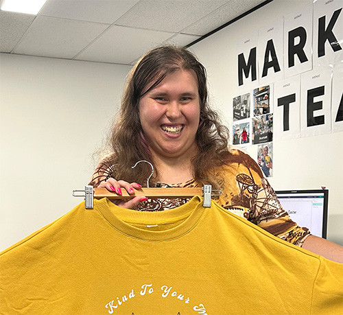 Amanda has long brown hair  and is smiling at the camera. She is holding up a yellow jumper. 