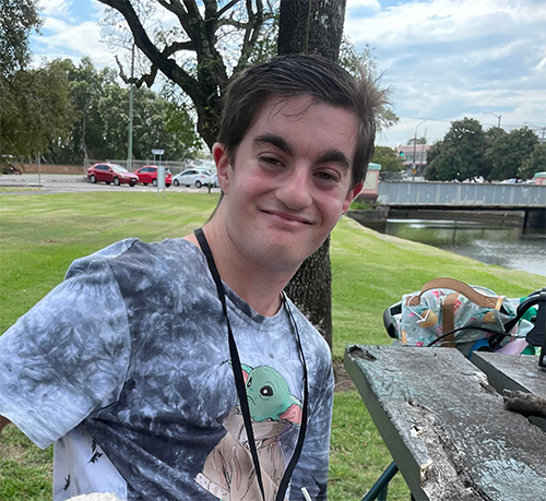 Joel is sitting in a park bench, he has short brown hair and wearing a grey t-shirt.