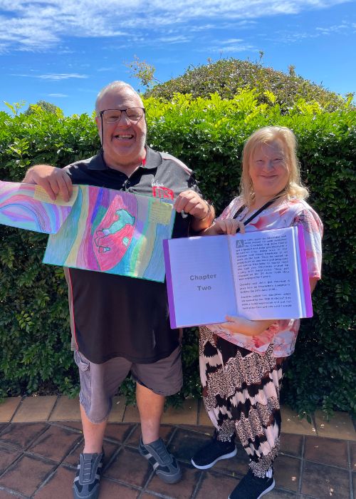 Mark and Maxine are standing in front of some green bushes. Maxine is holding her book. And Mark is holding his artwork. Both are smiling at camera.