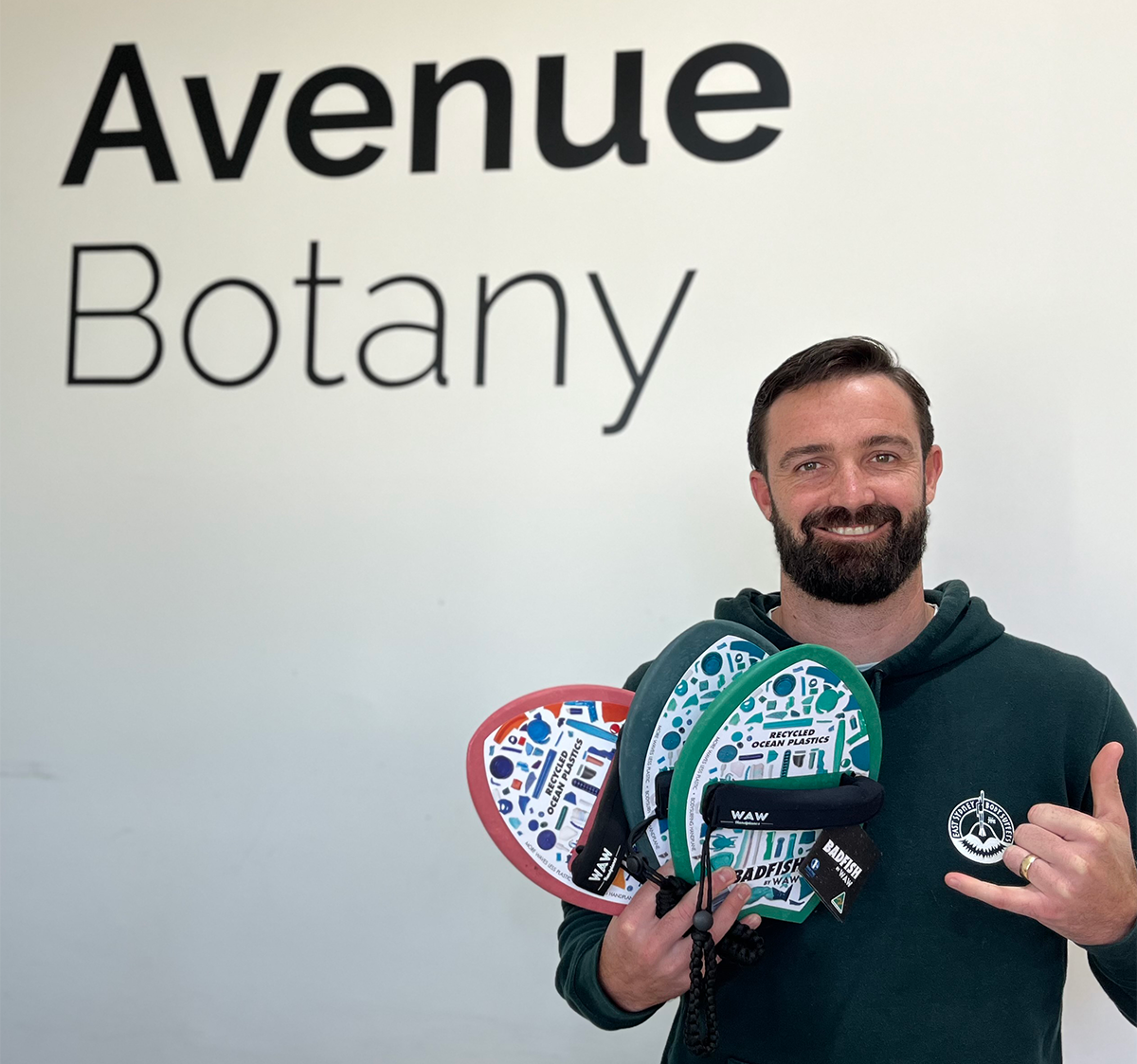 Rikki is standing in front of a white wall that says 'Avenue Botany'. He is holding three of his handplanes in his hands and is wearing a green jumper with his logo on it. He is smiling at the camera