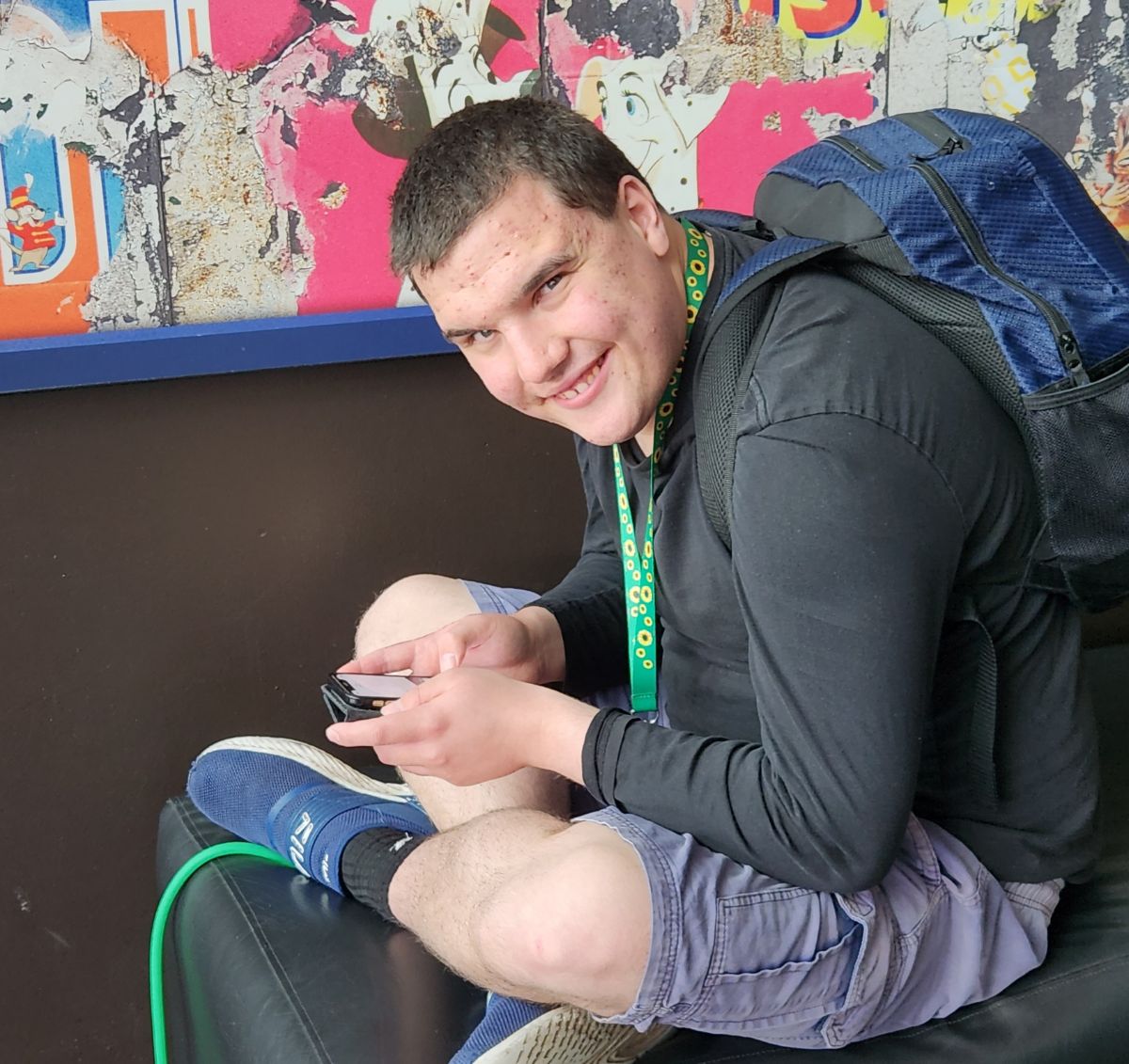 Robert is sitting on the floor, with his legs crossed smiling at the camera. He is wearing a blue backpack and a black jumper.