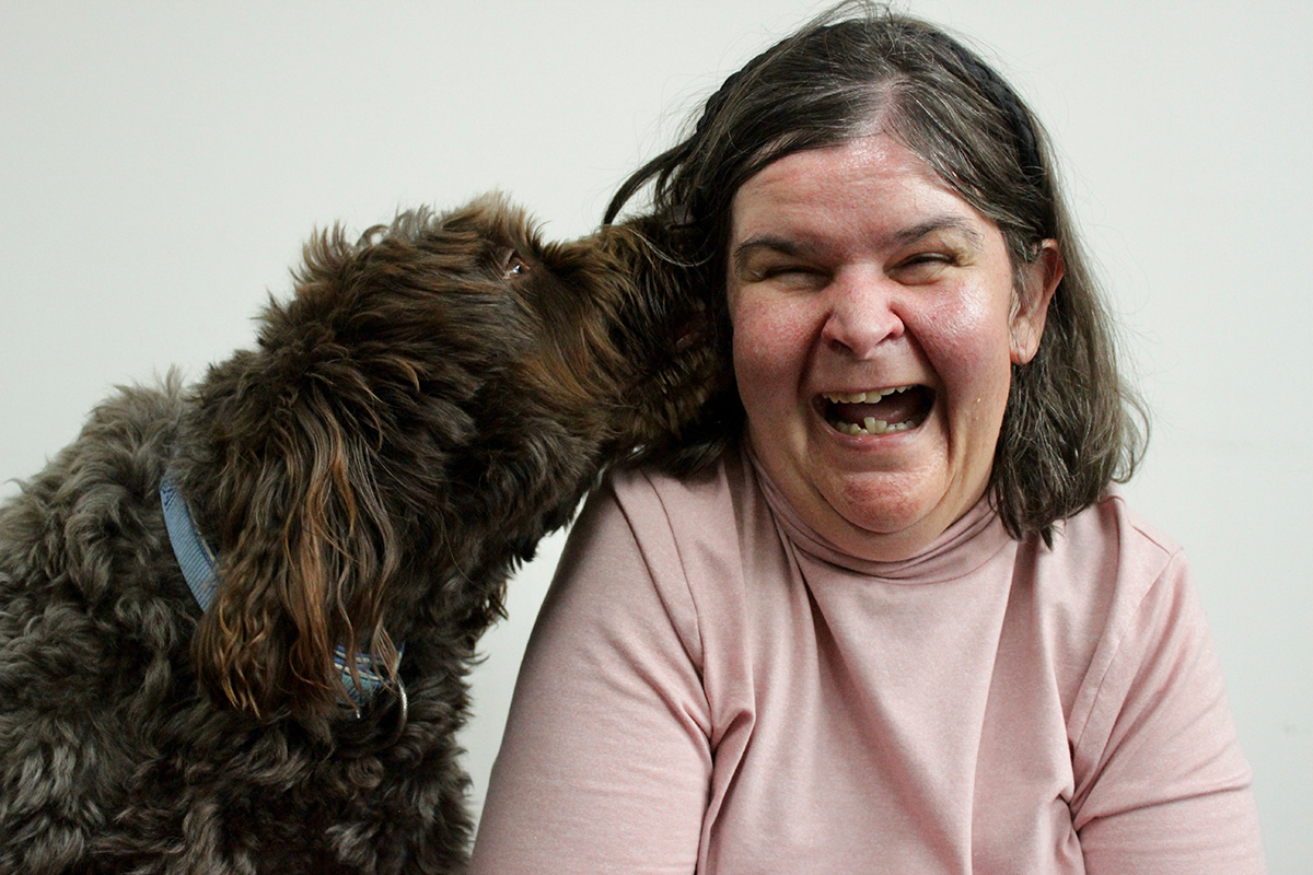 Avenue participant wearing a pink shirt with a brown dog that is kissing her cheek